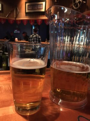 A glass and a pitcher of beer sit on a wooden bar counter in a dimly lit lounge.
