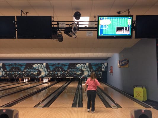 A woman in a pink shirt prepares to bowl on a lane with automatic scoring monitors above.