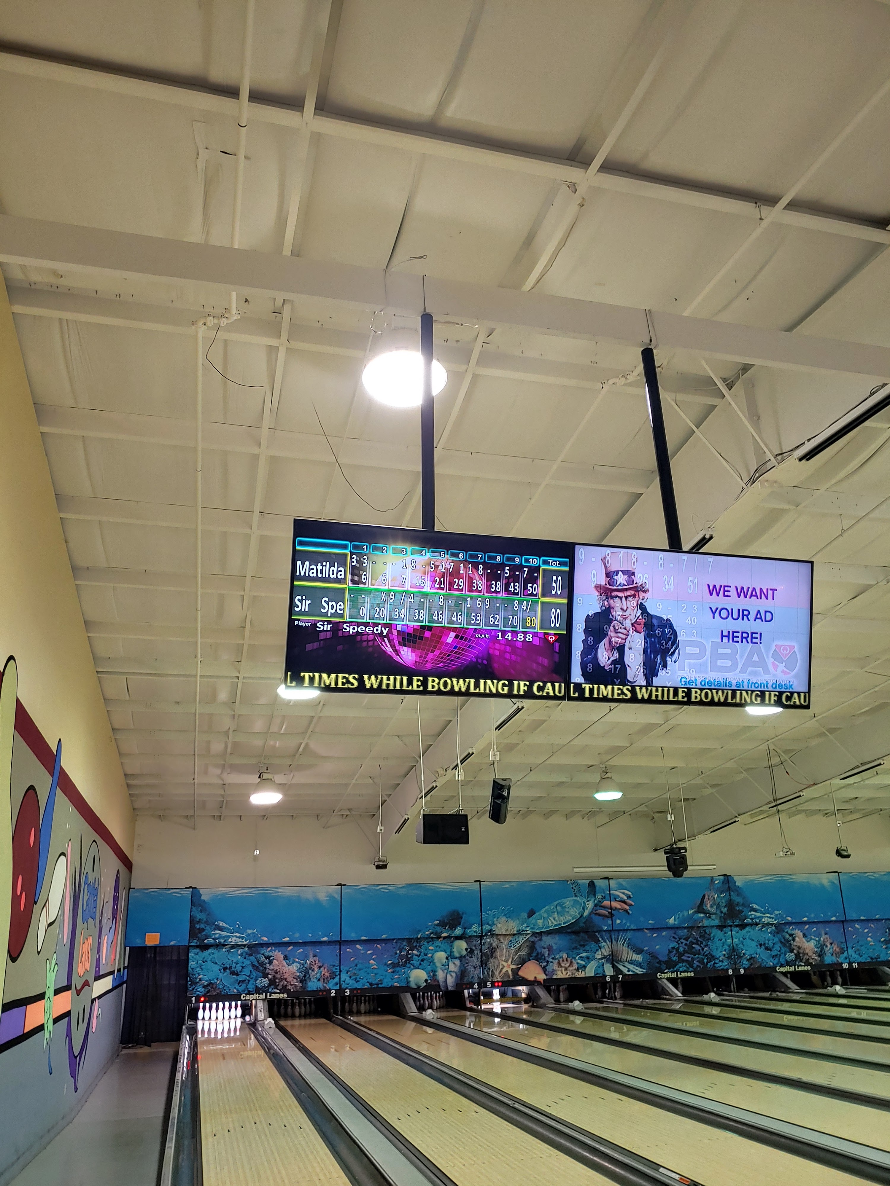A row of bowling lanes under bright lights with automatic scoring monitors and an underwater-themed mural backdrop.
