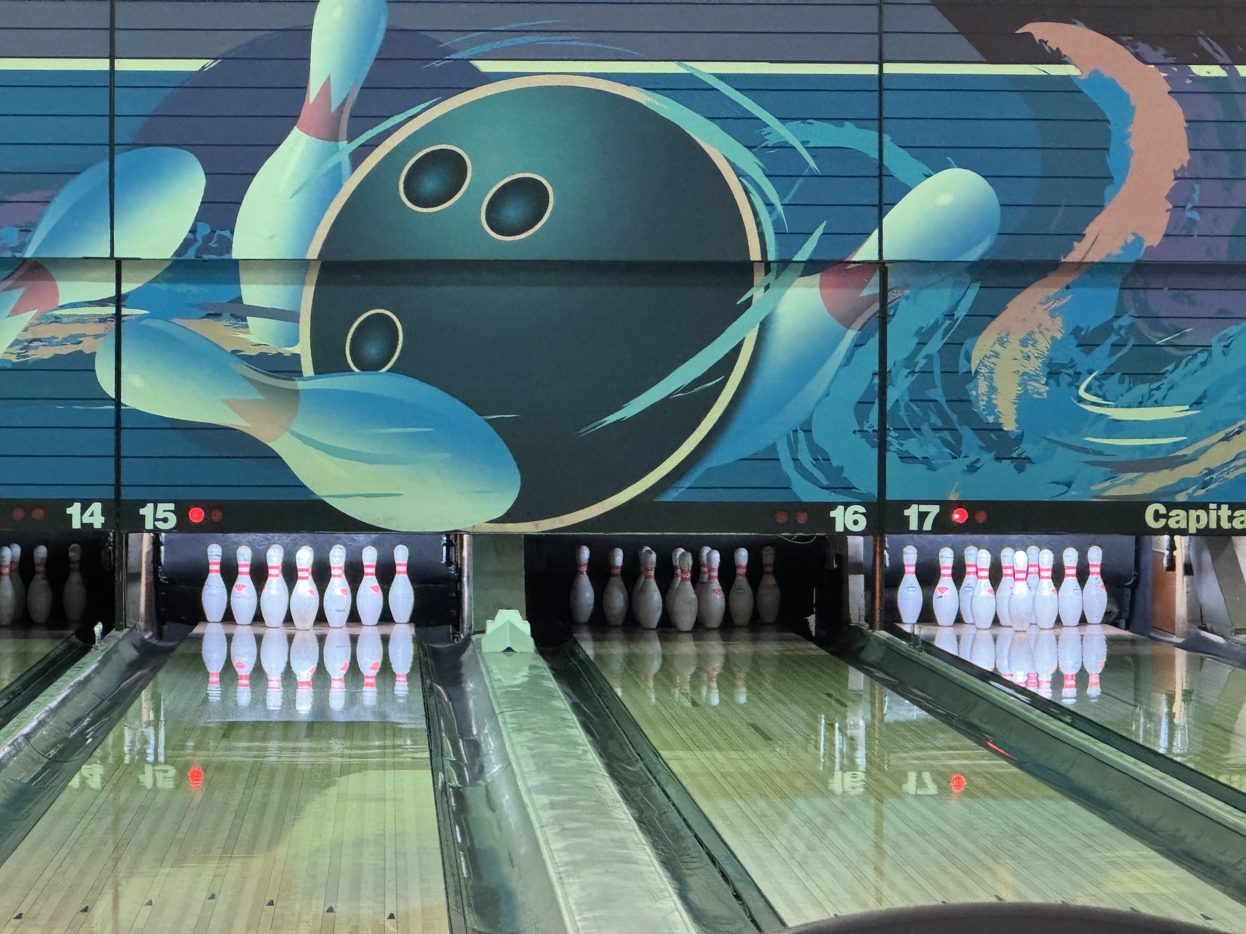 A straight-on view of four bowling lanes with a colorful mural of a bowling ball and pins on the back wall.