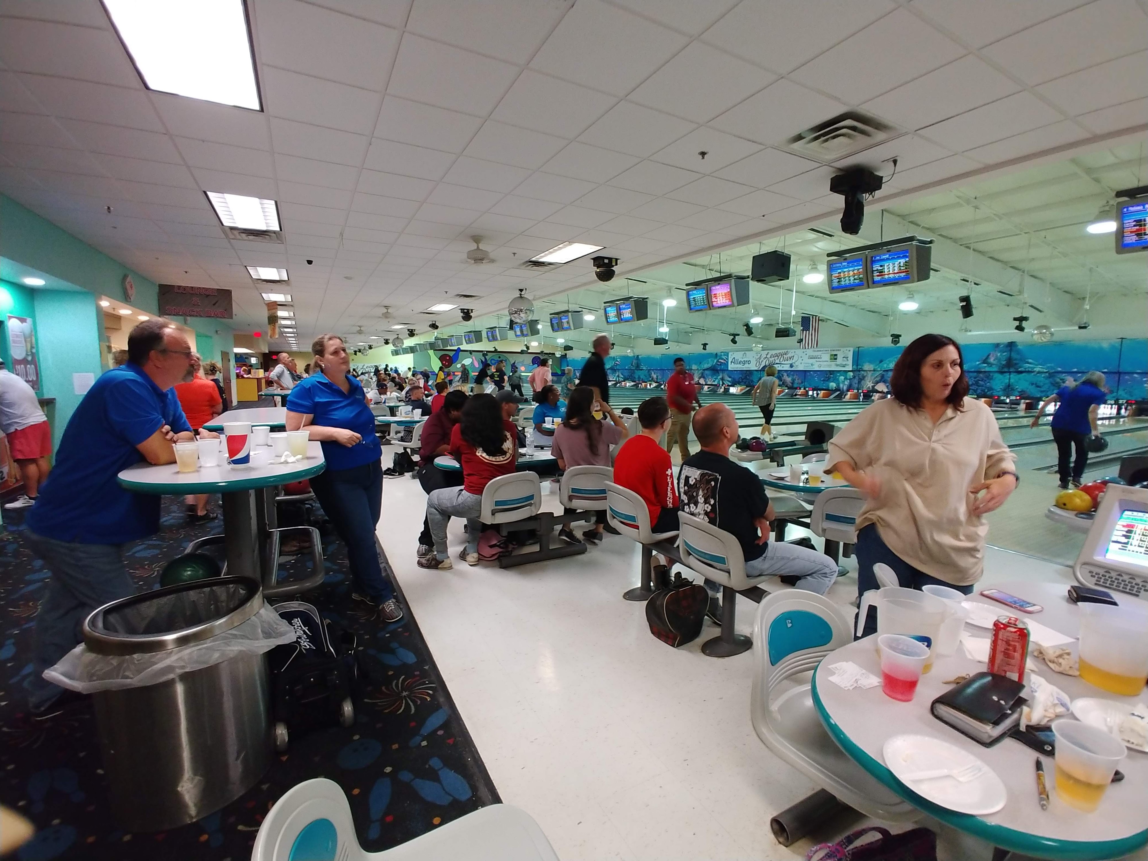 A busy bowling alley settee area with people sitting at tables and multiple lanes visible in the background.