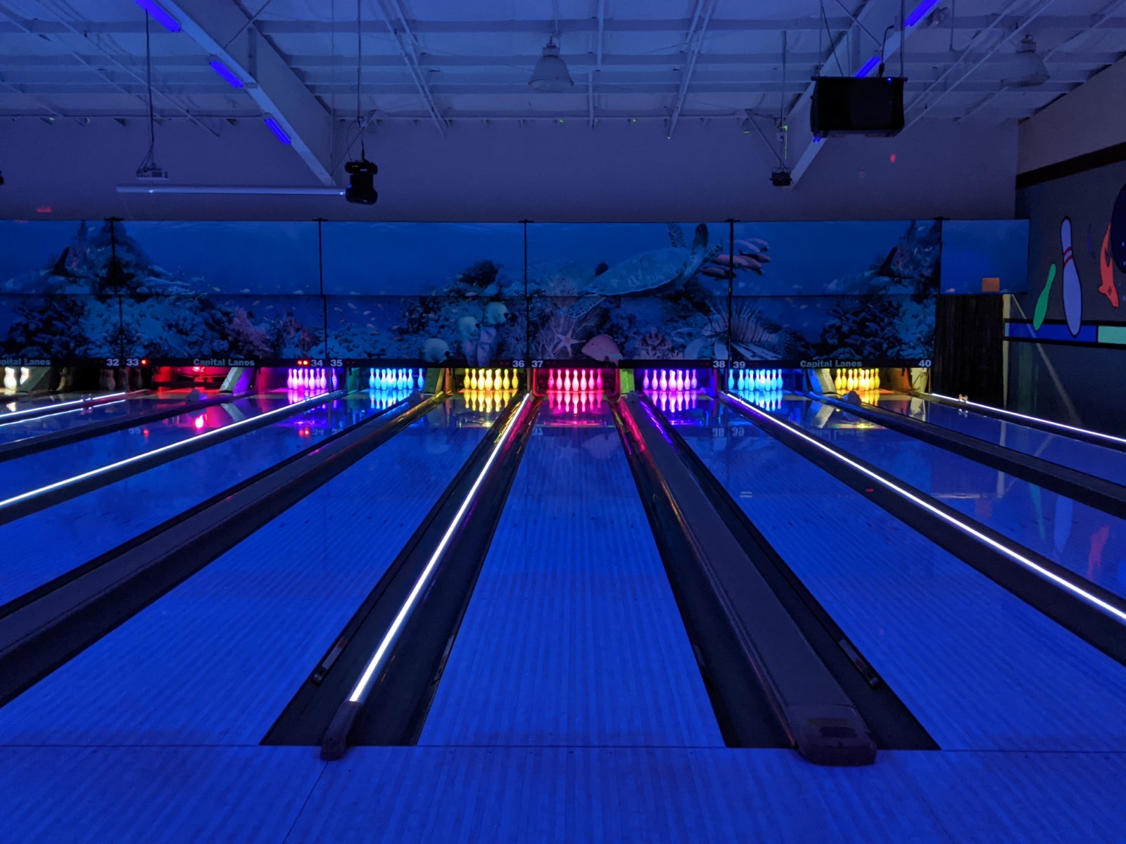 A symmetrical view of several bowling lanes under blue cosmic lighting with an underwater mural in the background.