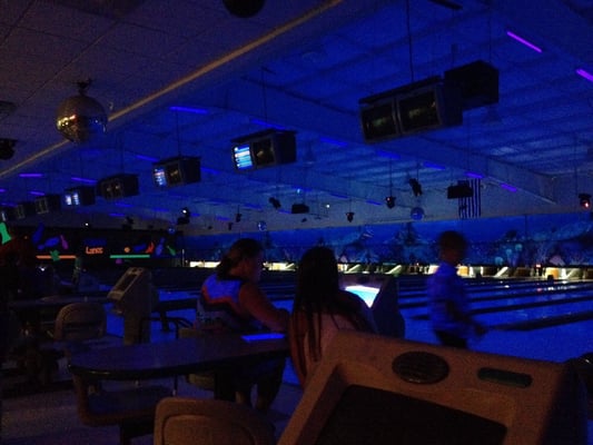 A bowling alley interior under vibrant blue cosmic lighting with lanes, scoring monitors, and people in silhouette.