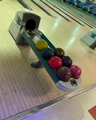 A bowling ball return rack filled with colorful bowling balls on a wood-grain lane.