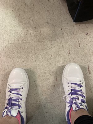 Top-down view of a person wearing white bowling shoes with purple laces on a tiled floor.