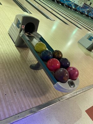 A bowling ball return rack filled with several colorful bowling balls on a wooden lane.