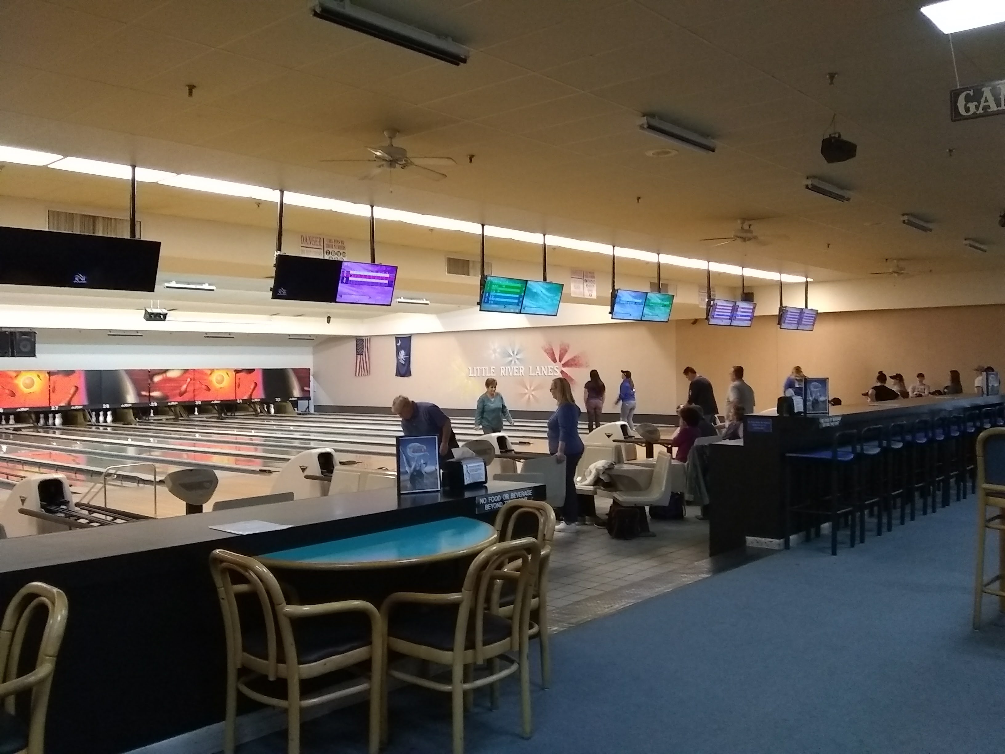 Interior view of Little River Lanes featuring several bowling lanes, overhead scoring monitors, and people socializing.