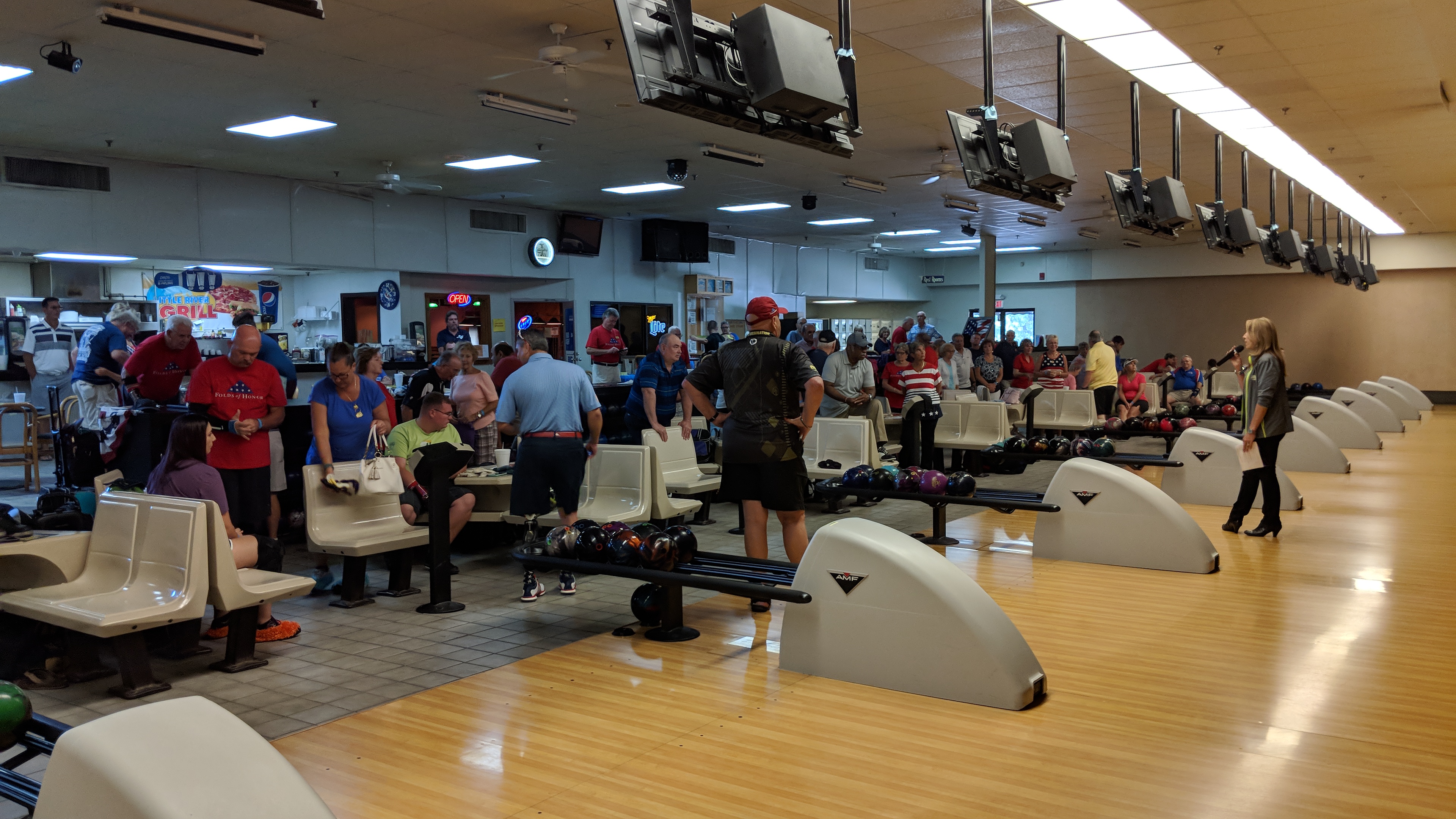 A crowded bowling alley where a woman with a microphone addresses a large group of people near the lanes.