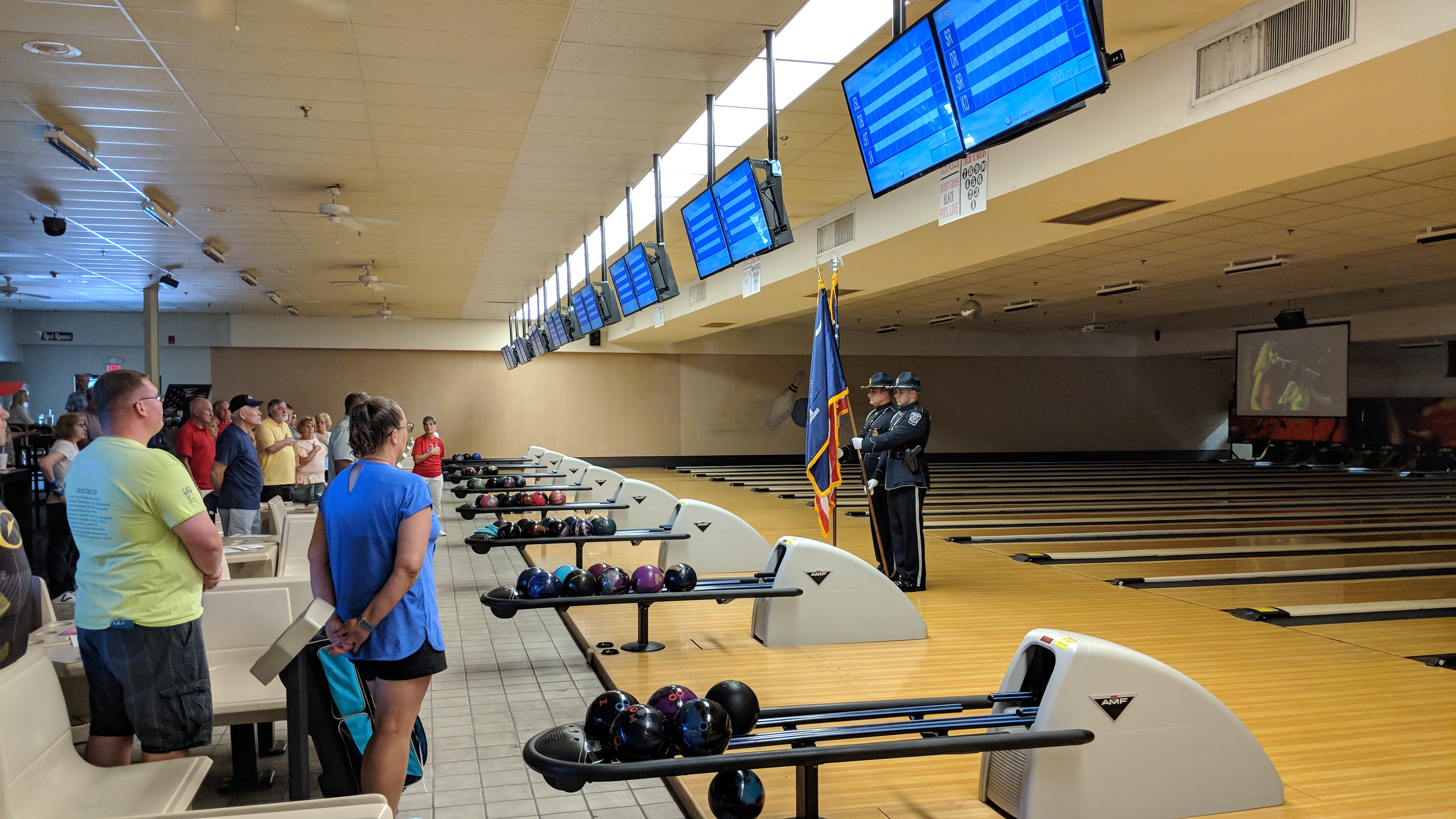 Two uniformed officers hold flags on bowling lanes in front of a standing crowd.