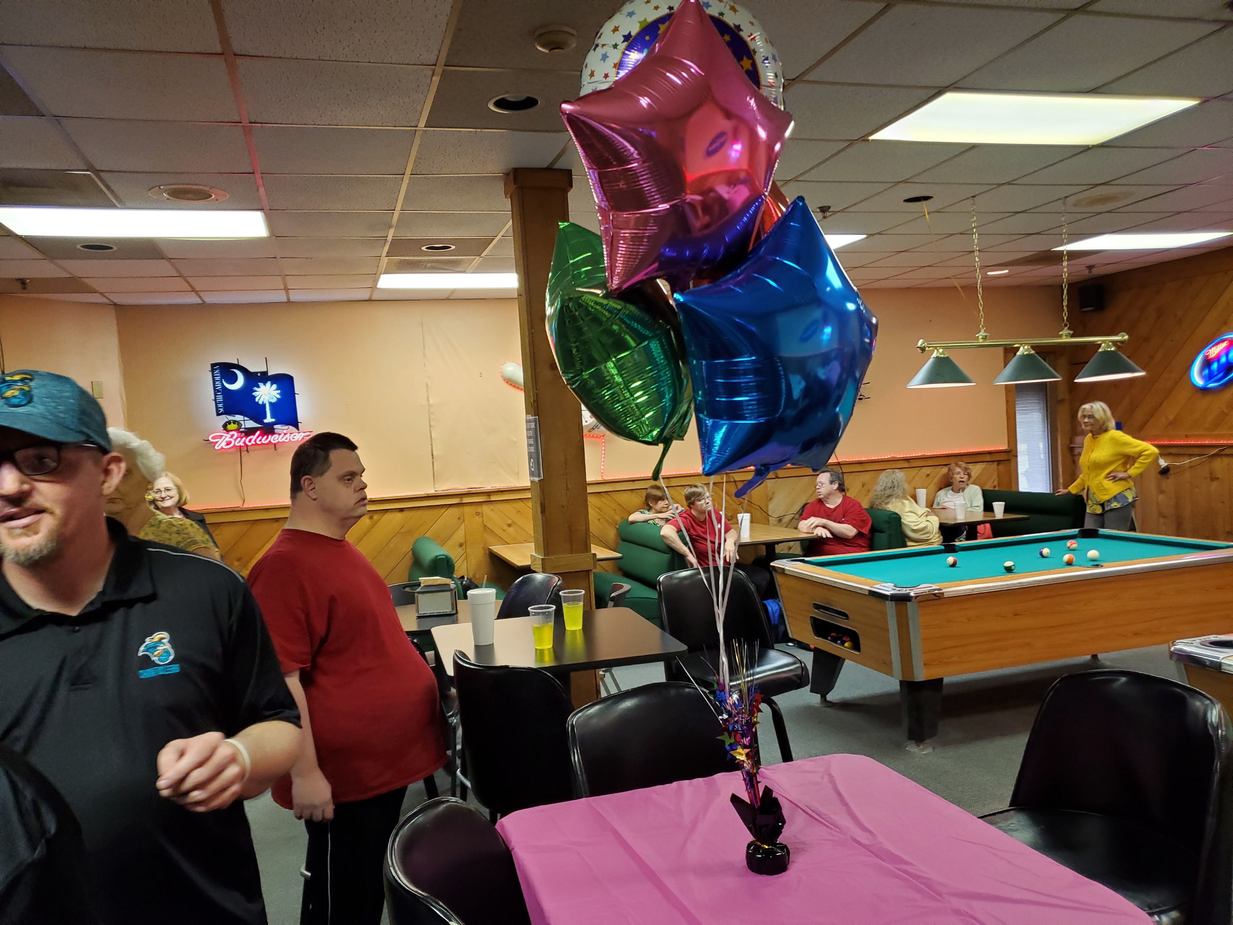 A festive lounge area with star balloons on a pink table and people gathered around a billiards table.