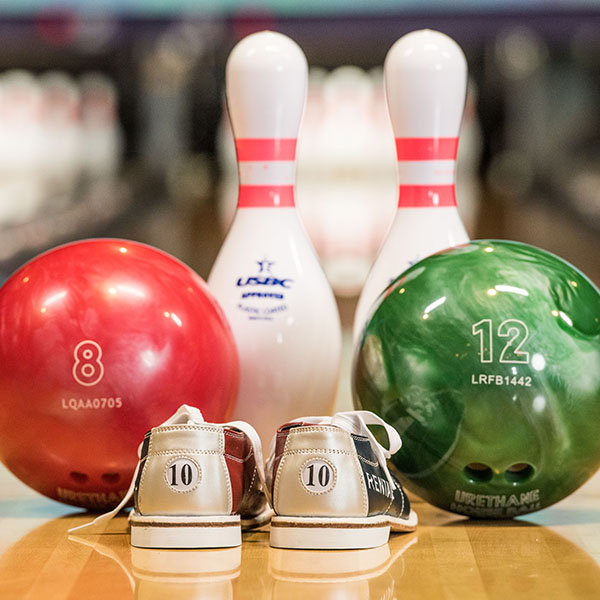 A red bowling ball, a green bowling ball, two pins, and rental shoes sitting on a bowling lane.
