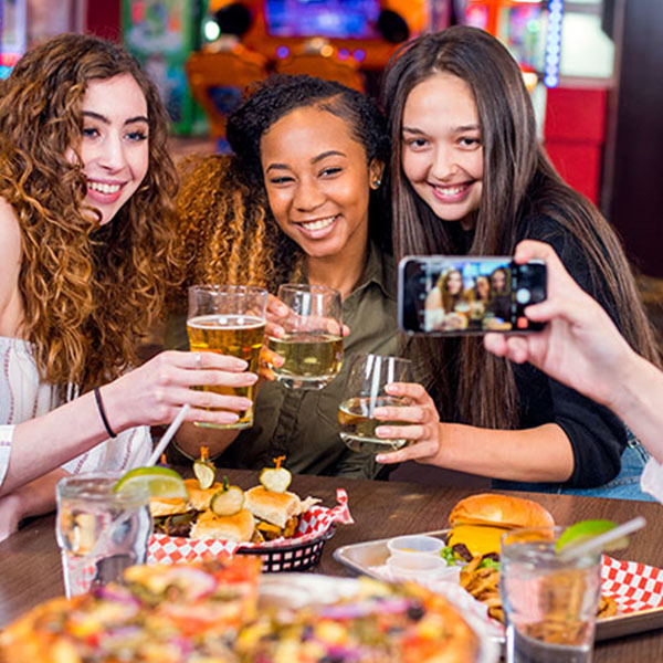 Three smiling friends holding drinks and taking a selfie over a table filled with pizza, sliders, and burgers.