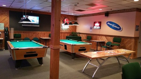 Two green billiards tables in a wood-paneled lounge area with chairs and neon signs.
