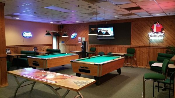 A pool room with two green felt billiard tables, neon beer signs, and green seating booths.