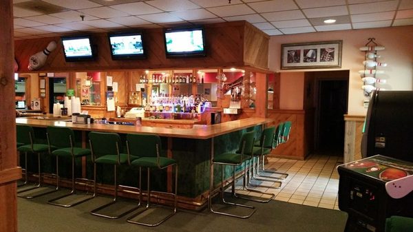 A wooden bar with green stools and three overhead televisions at Little River Lanes.