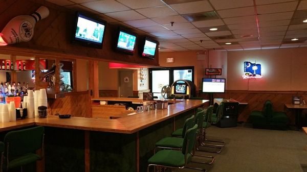 A wooden L-shaped bar with green stools, wall-mounted televisions, and a neon sign in a lounge area.