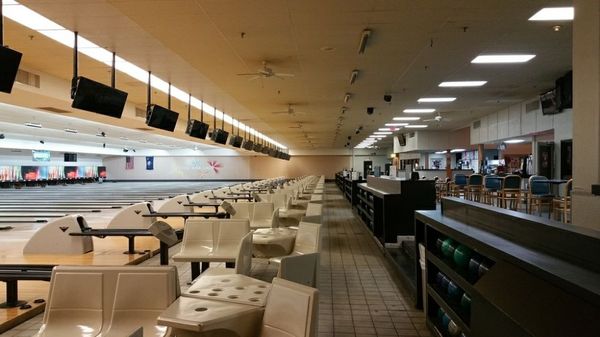 A wide view of a bowling alley with white seating, several lanes, and a bar area in the background.