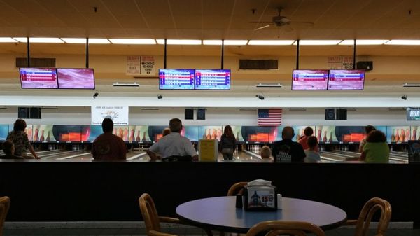 People watch from a seating area as others play on several bowling lanes with digital score screens overhead.