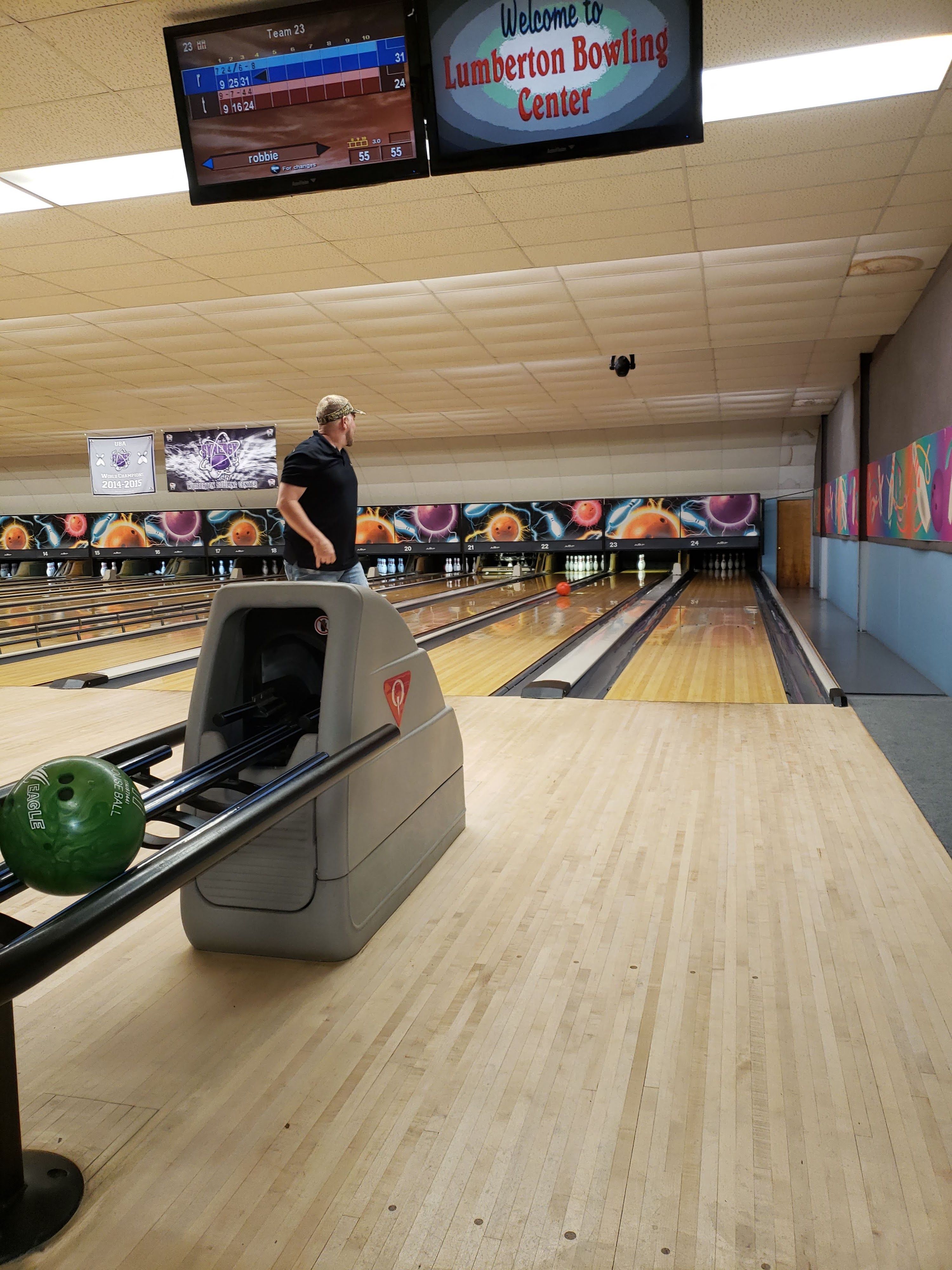 A man watches his ball roll down a lane at Lumberton Bowling Center with overhead score monitors visible.