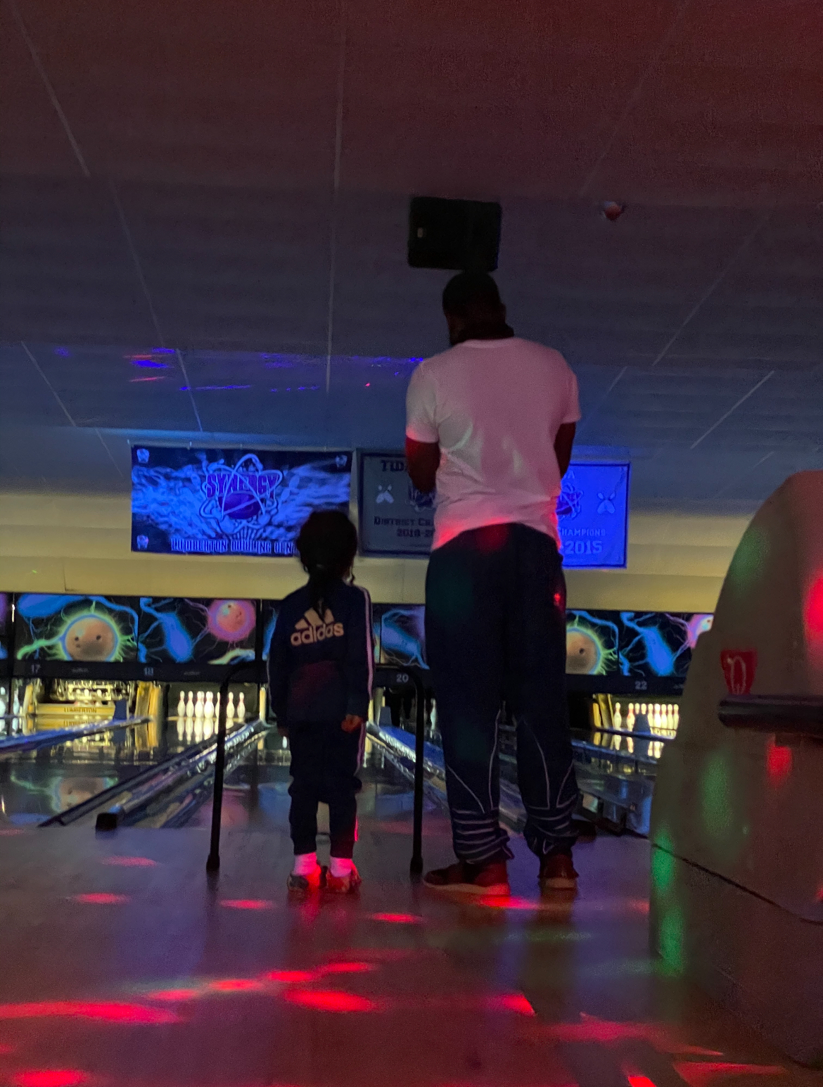 A man and a young child stand at the start of a bowling lane under colorful cosmic lights.