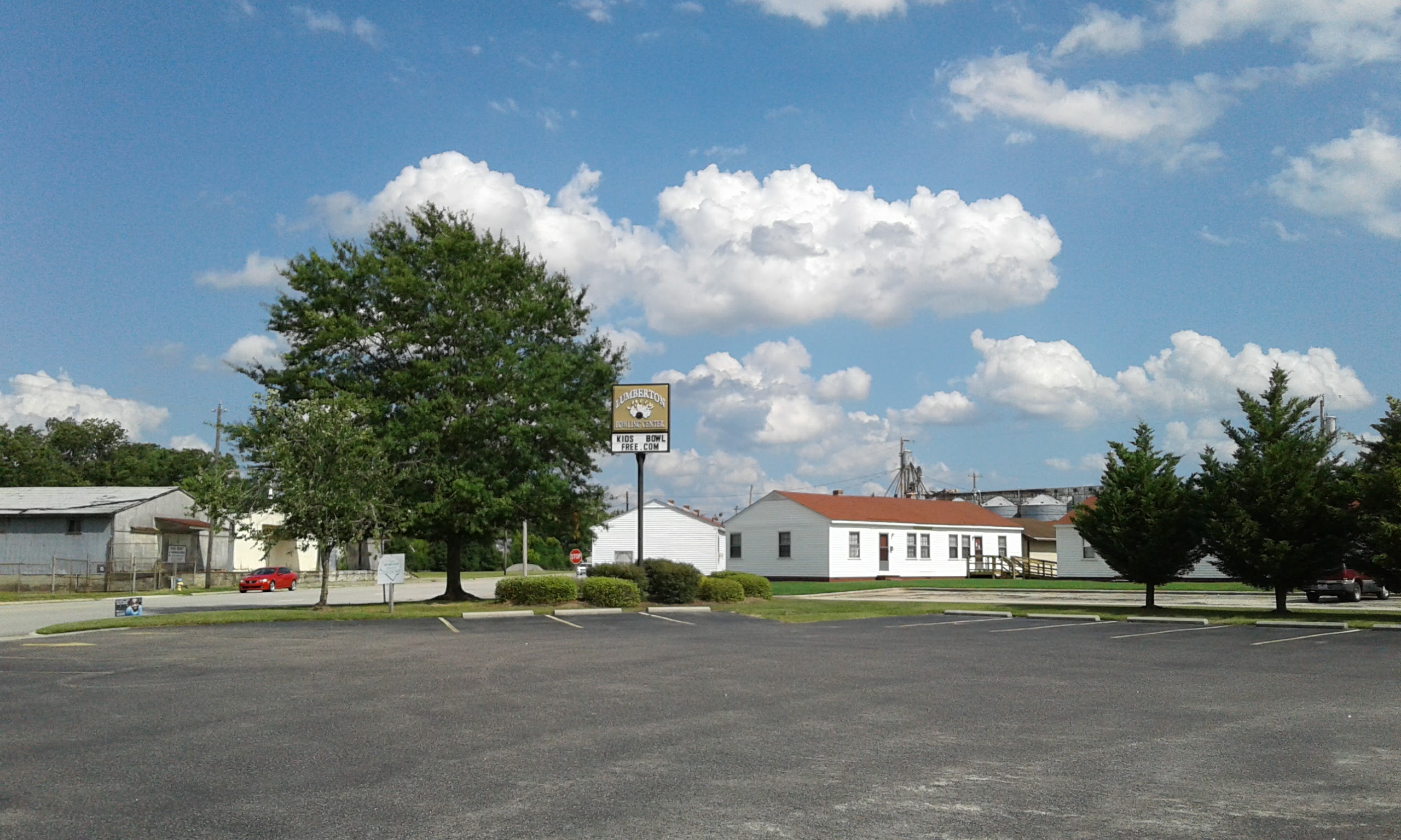 An outdoor photo of a large asphalt parking lot with a sign for Lumberton Bowling Center, surrounded by trees and buildings under a blue sky.