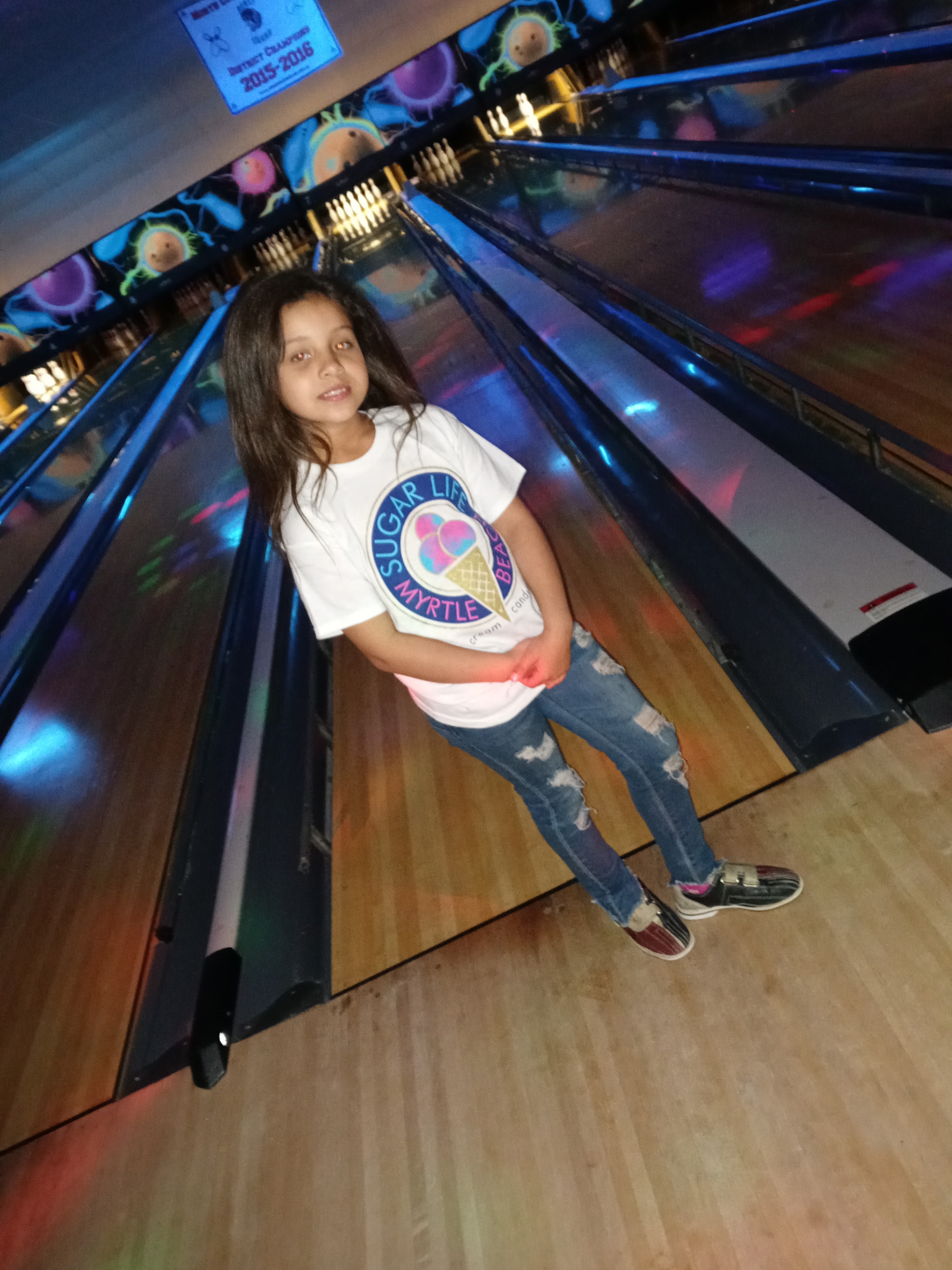 A young girl smiles while standing on a bowling lane approach during cosmic bowling, wearing bowling shoes.