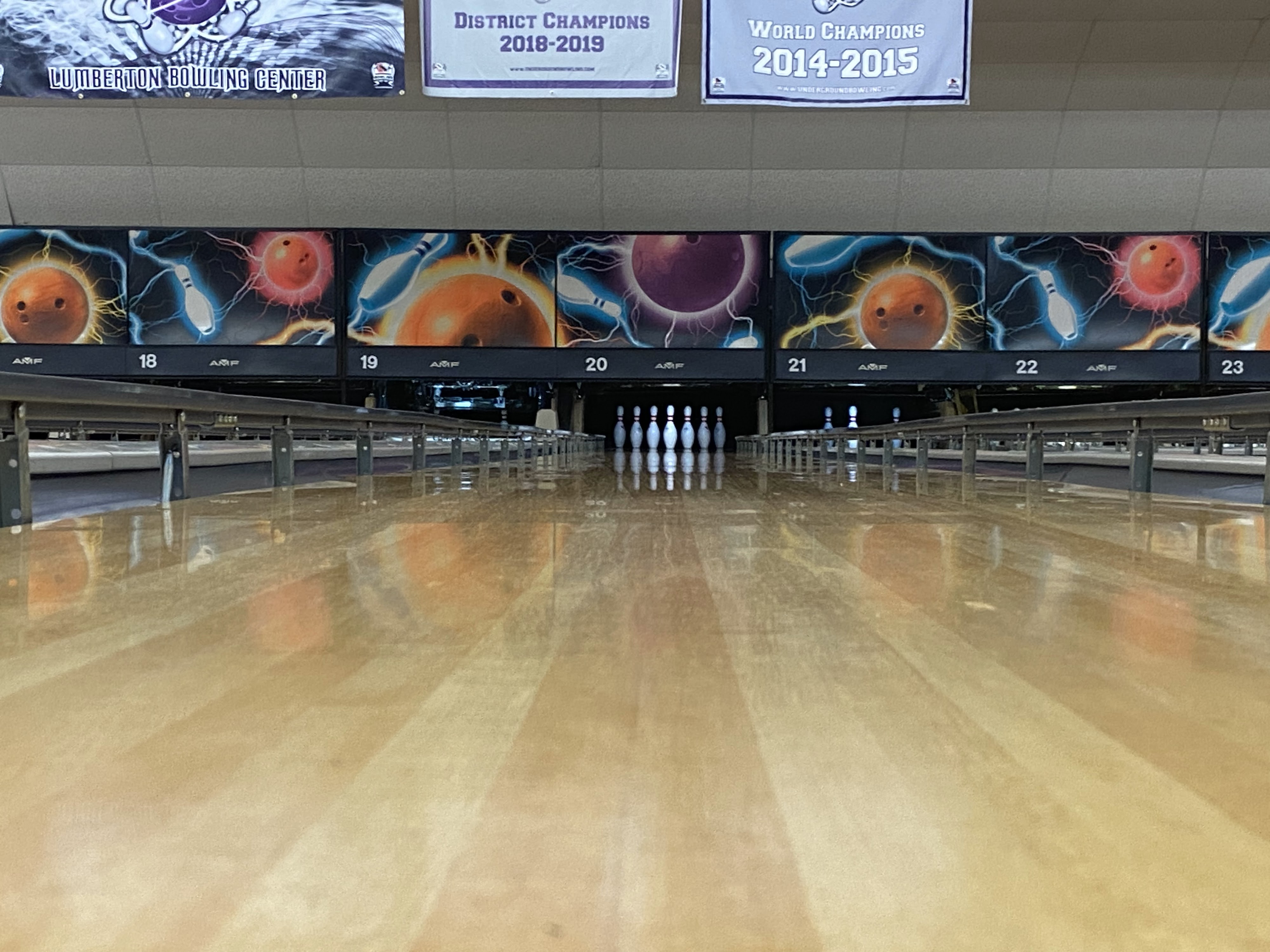 A bowling lane with a full rack of white pins at the end, flanked by digital scoring displays and championship banners.