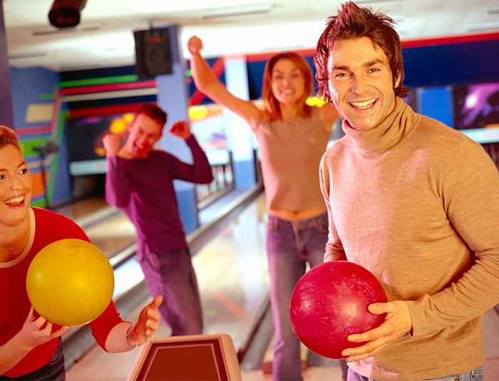 Four smiling adults bowling, with two holding bowling balls and two celebrating in the background.