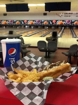 A sandwich and crinkle-cut fries in a checkered basket with a Pepsi cup in front of bowling lanes.