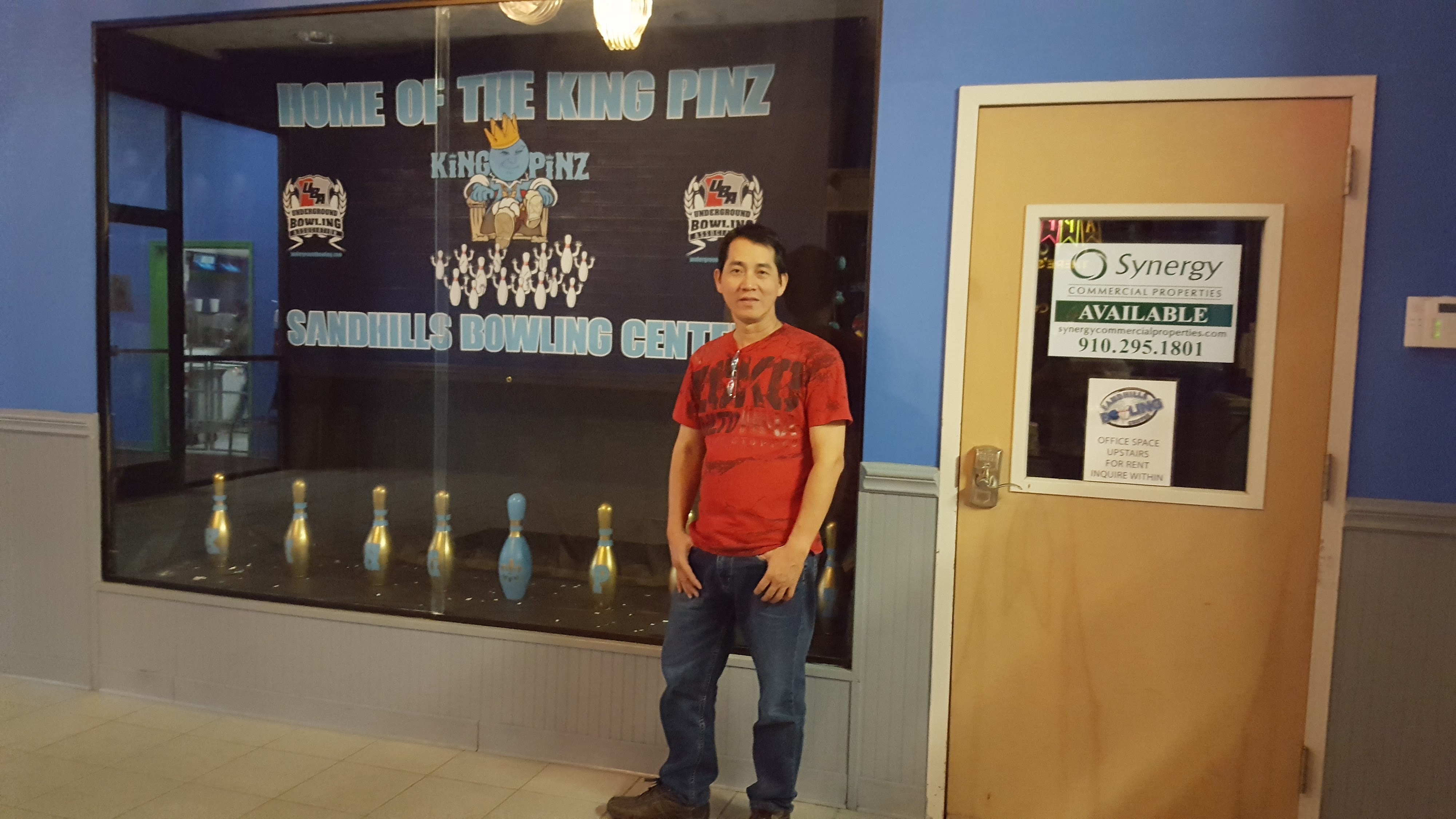 A man in a red shirt stands in front of a bowling display case with gold and blue pins and league logos.
