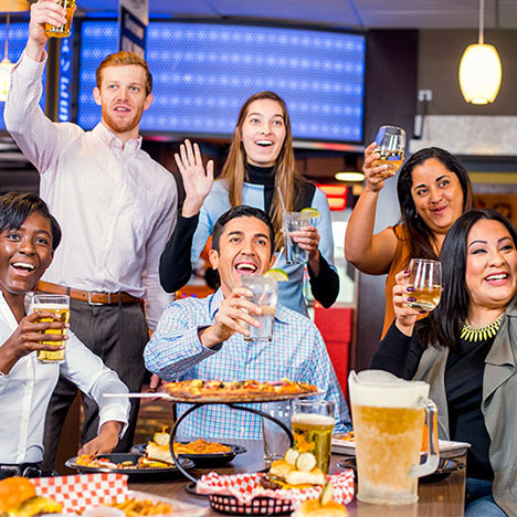 A group of diverse friends smiling and raising their glasses in a toast over a table filled with pizza and burgers.