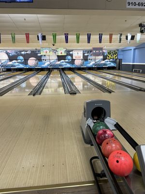 A row of colorful bowling balls sits in a ball return looking down clean, empty bowling lanes.
