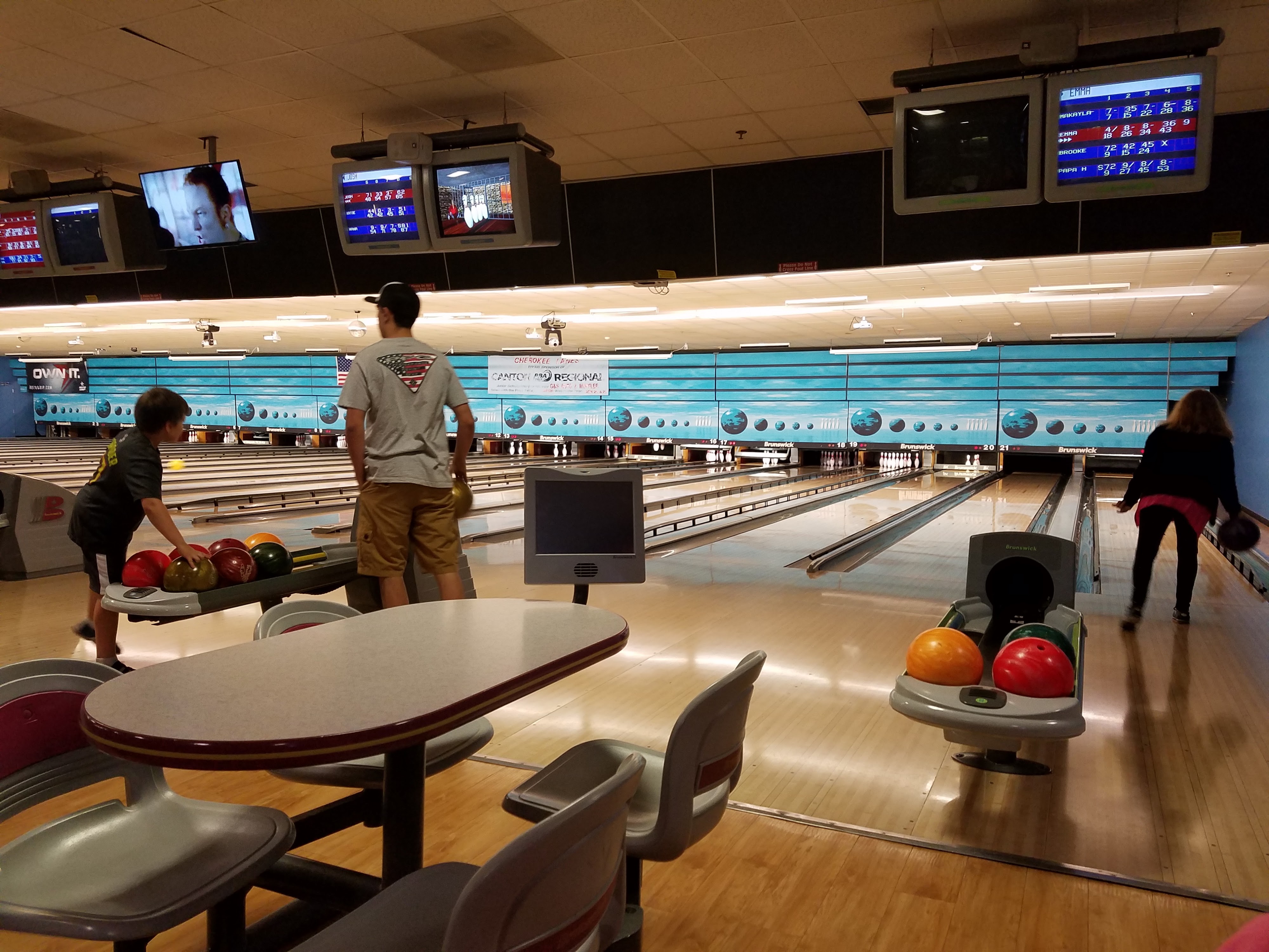 People bowling at an indoor center with blue wall graphics and overhead scoring monitors.