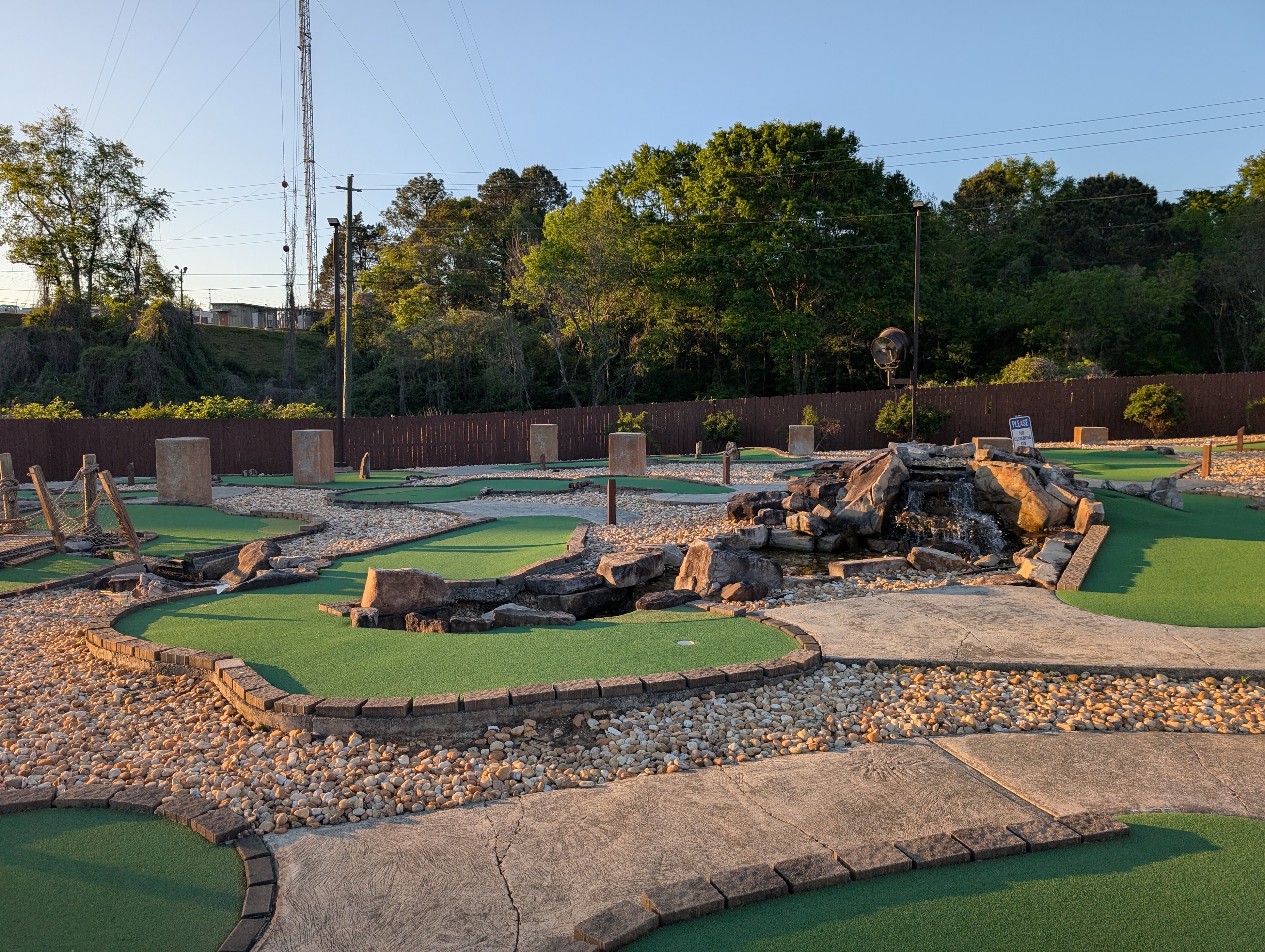 An outdoor miniature golf course with green turf, stone paths, and a rock waterfall feature under a clear sky.