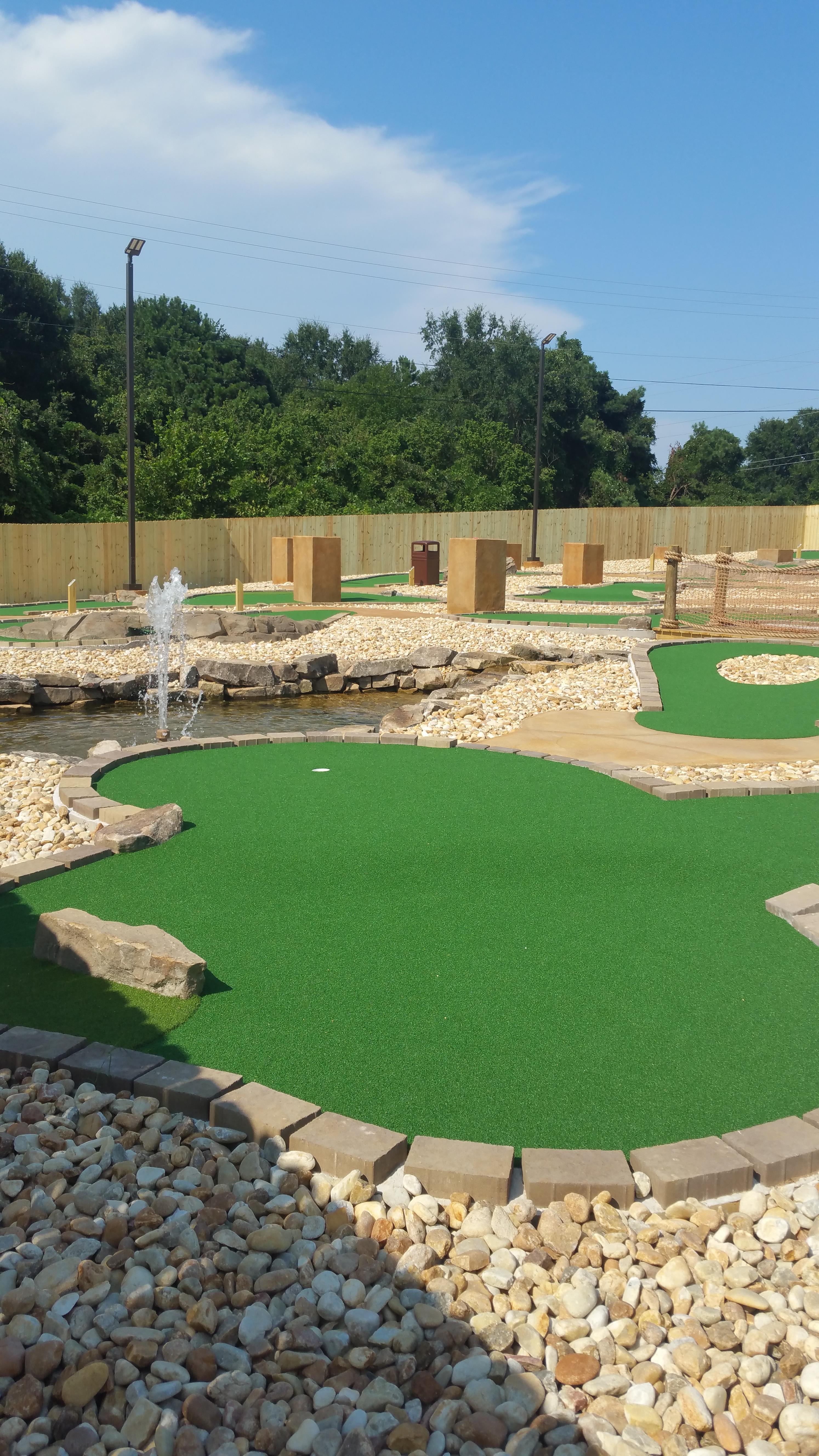 An outdoor mini-golf course with bright green turf, stone borders, and a small water fountain under a blue sky.