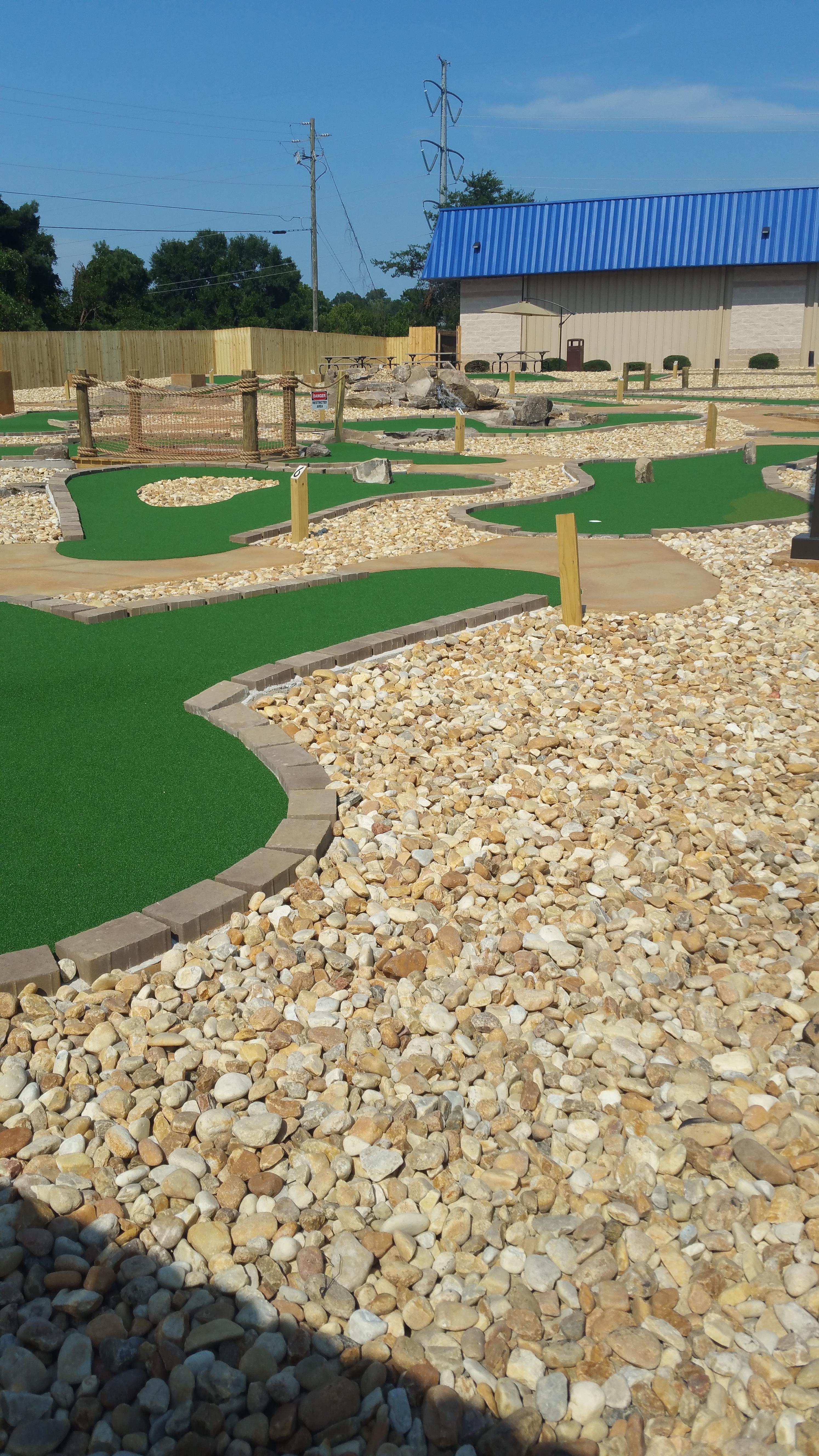 An outdoor mini golf course with green turf and stone pathways under a blue sky.