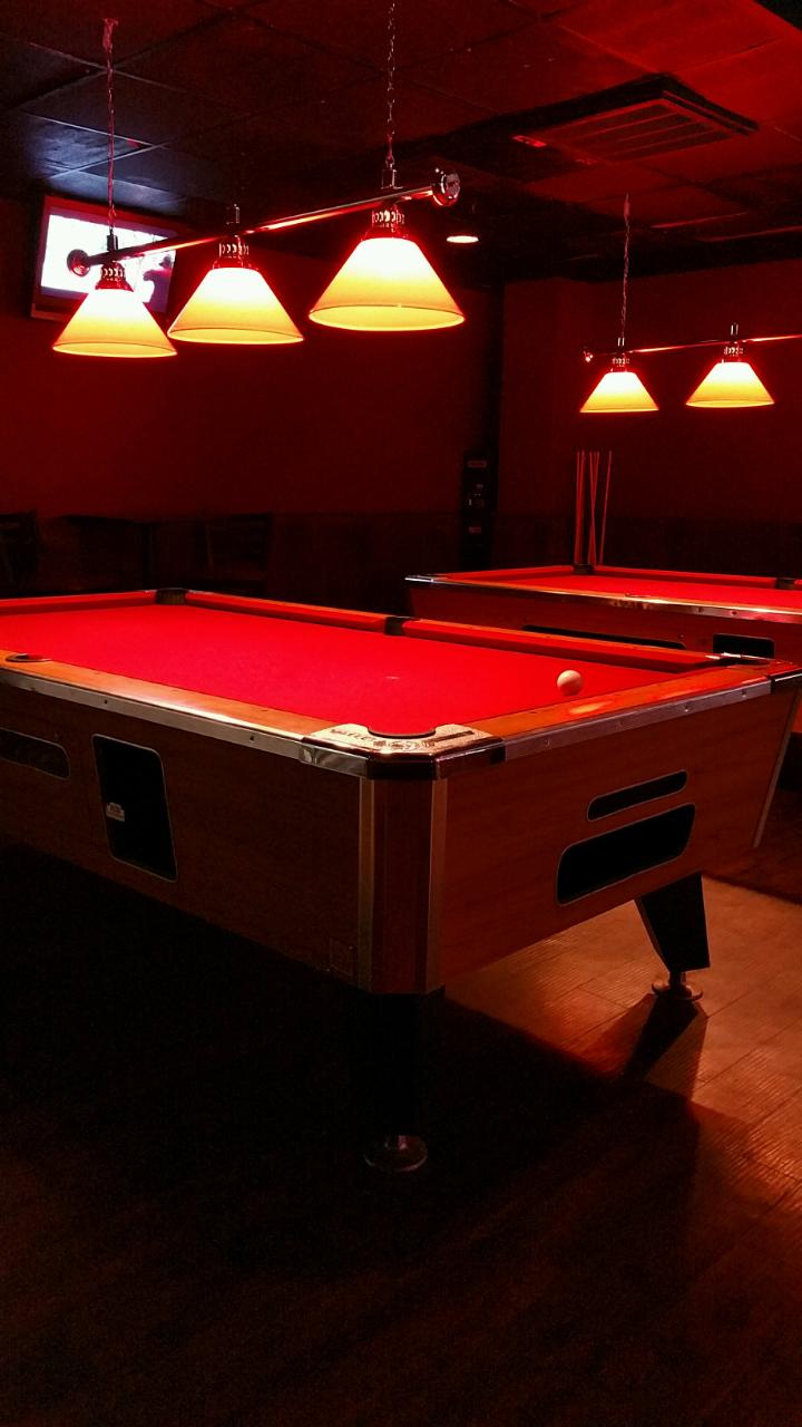 Two billiards tables with red felt illuminated by warm pendant lights in a dark room.
