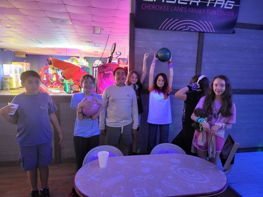 A group of happy children holding bowling balls under purple cosmic lighting in a bowling center settee area.