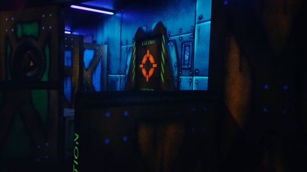An indoor laser tag arena with neon painted walls, metallic textures, and a glowing orange target under blacklights.