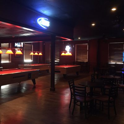 A dimly lit lounge featuring several pool tables with red felt and small dining tables.
