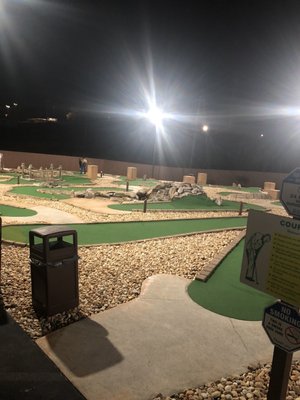 An outdoor mini golf course at night featuring green turf, rock obstacles, and bright overhead lights.