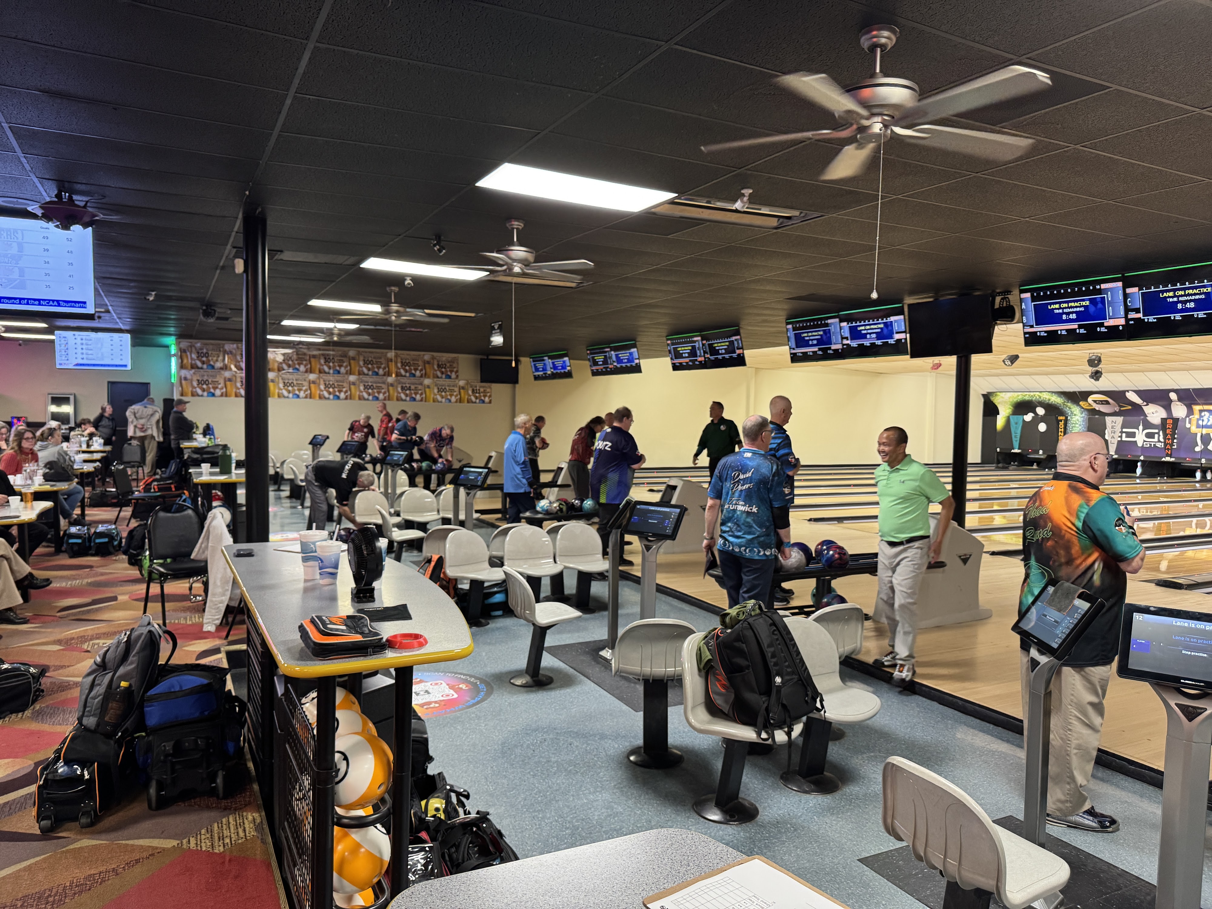 Bowlers in jerseys stand near the lanes and automatic scoring monitors inside a well-lit bowling center.