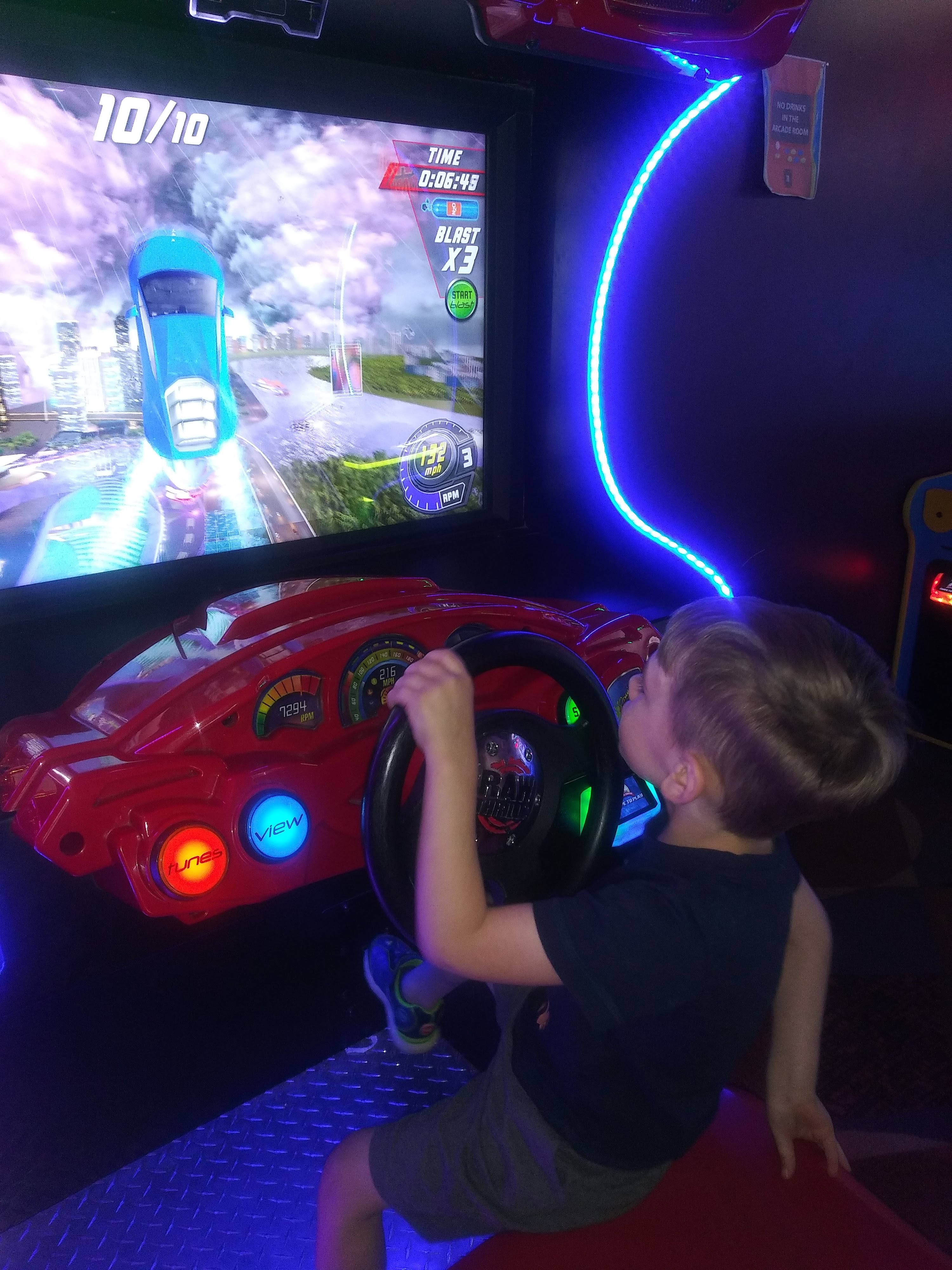 A young boy concentrates while playing a car racing simulator in an arcade with blue neon lights.