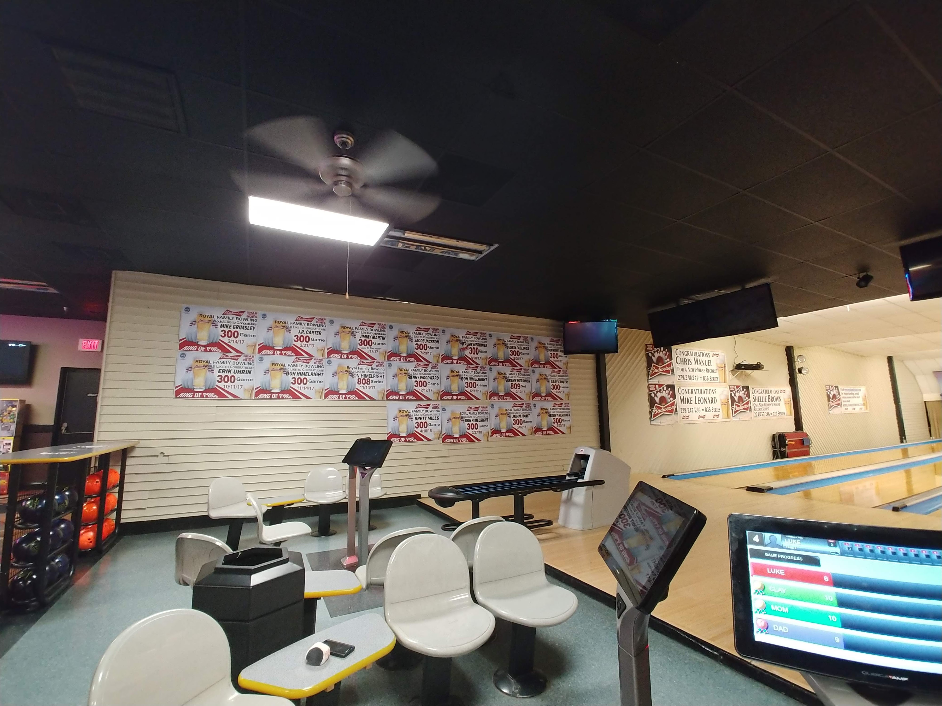 A bowling center settee area featuring white chairs, scoring tablets, and a wall of 300-game achievement posters.