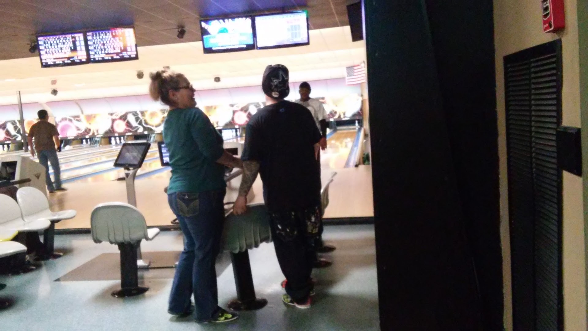 People socialize near the lanes in a bowling alley with digital scoring monitors overhead.