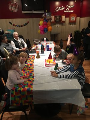 Children and adults wearing party hats sit at a decorated table with a birthday cake in the Strike Zone Lounge.