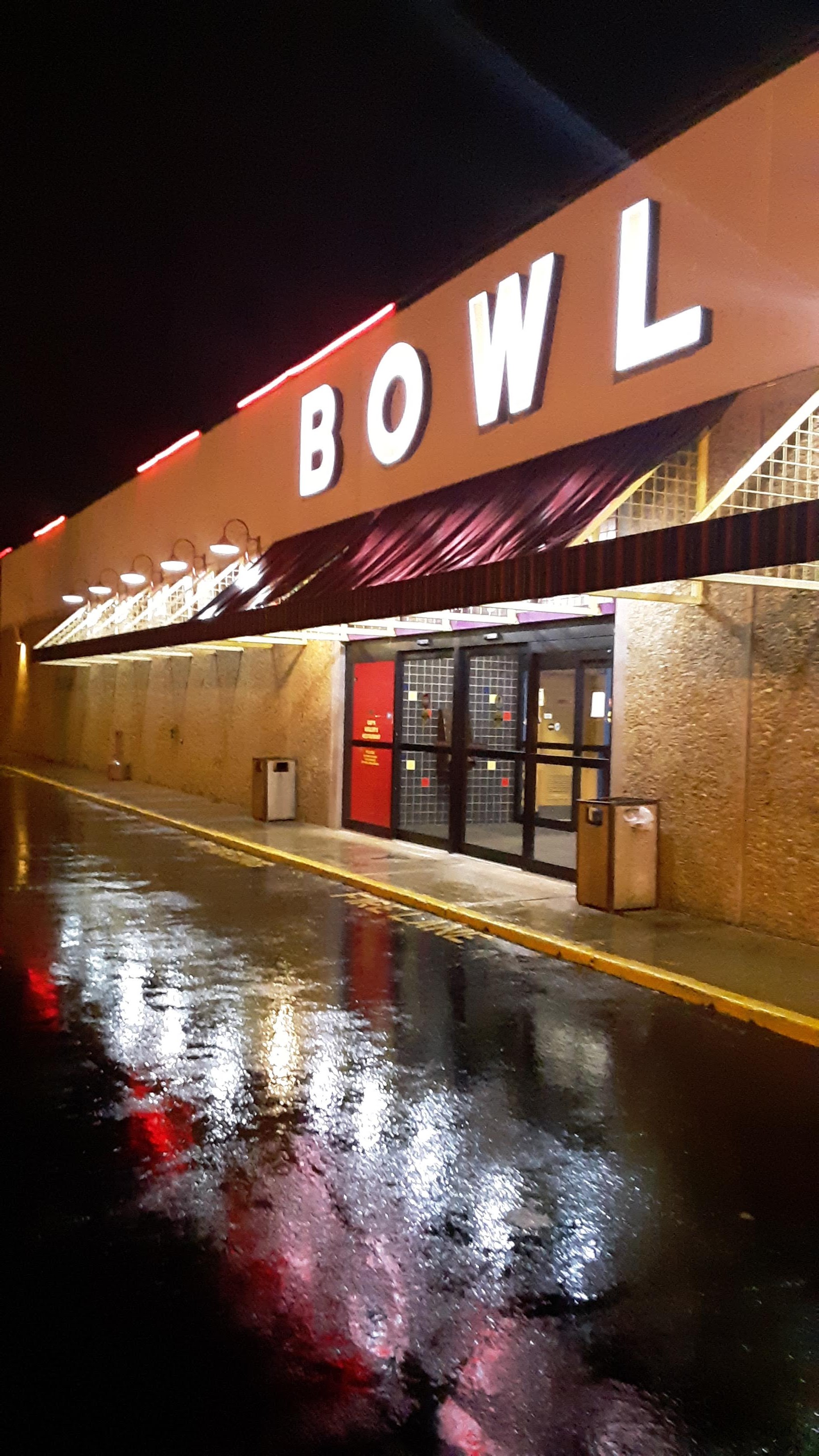 Nighttime exterior of a bowling center with glowing white signage and red neon reflections on wet asphalt.