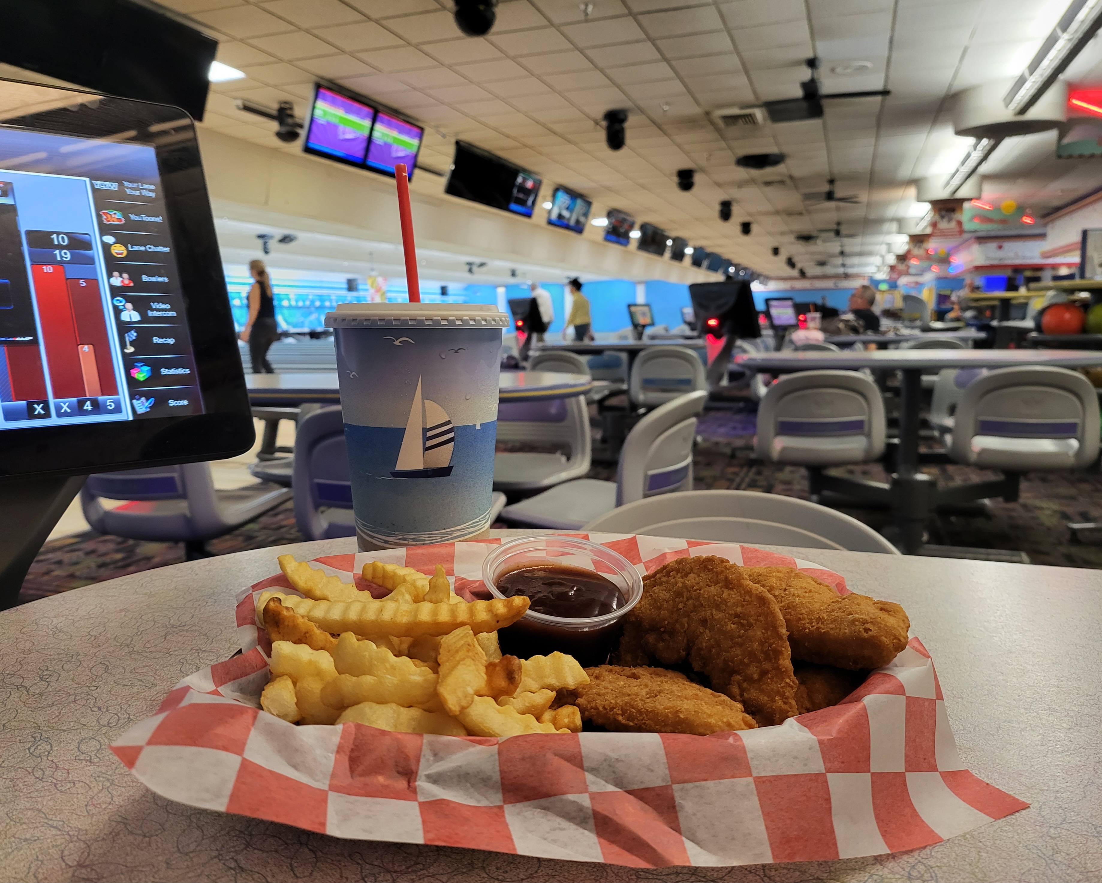 A basket of chicken tenders and fries on a table next to a soft drink and a bowling scoring tablet.