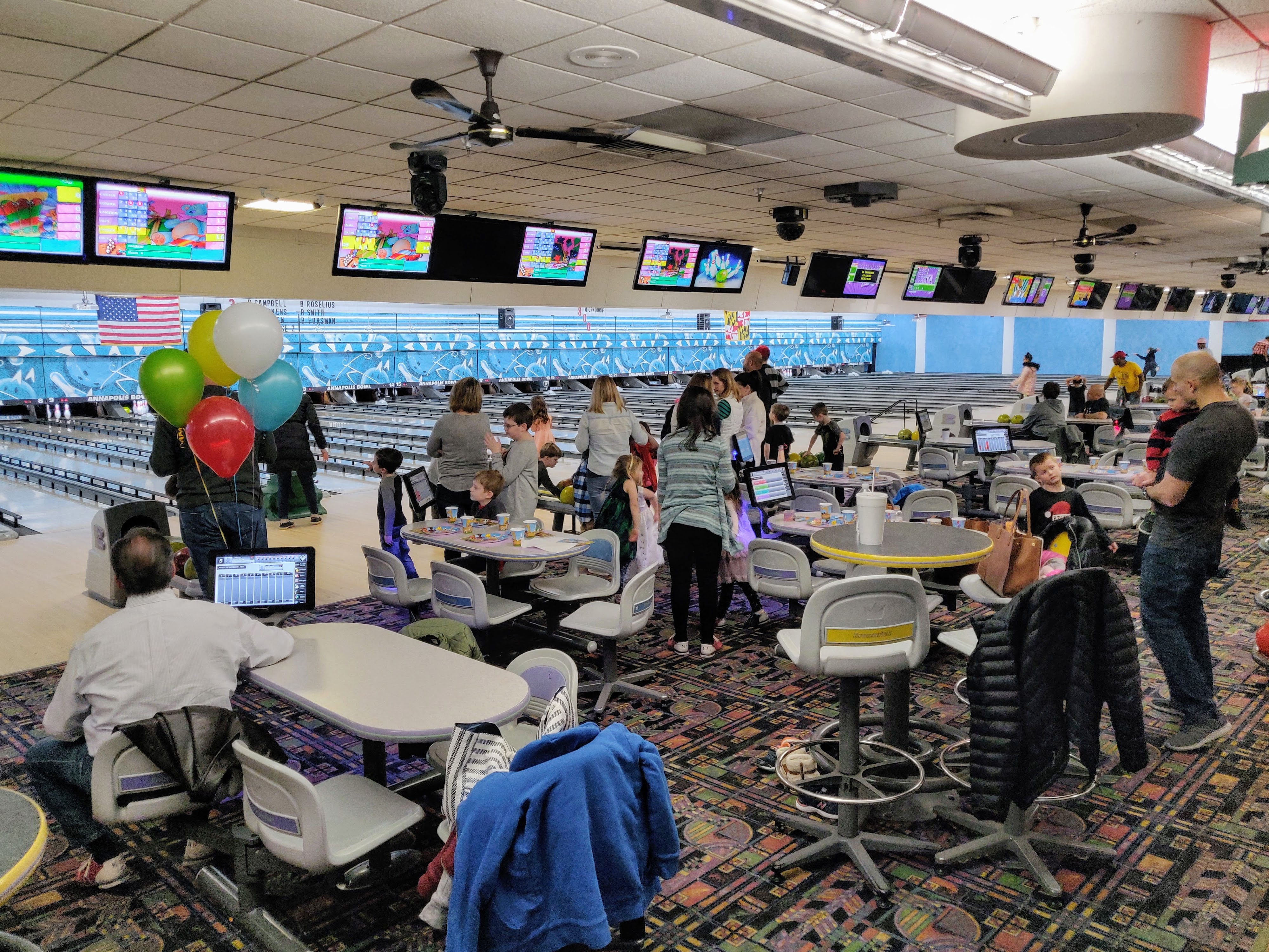 A group of families gathered for a party at a bowling alley with colorful balloons and lanes in the background.
