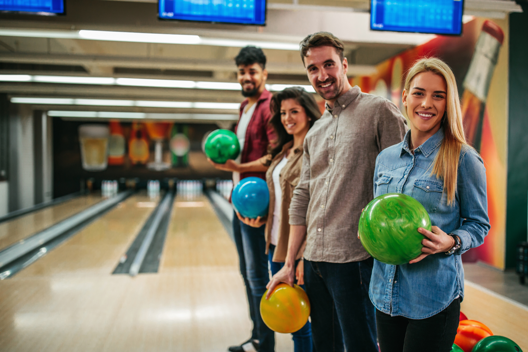 Four smiling friends standing on a bowling lane holding colorful bowling balls.